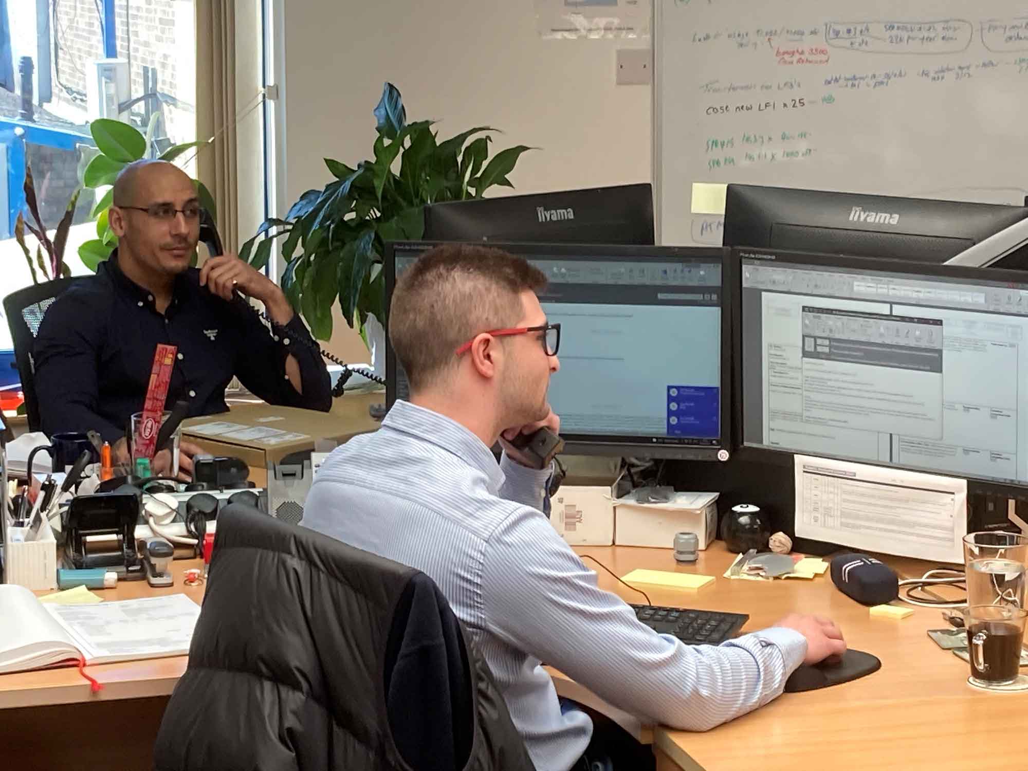 Two men in an office sat at their desks while holding a phone to their ear to help illustrate GB Electronics' after sales service.